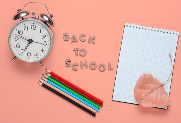 Notepad, colored pencils, alarm clock on pink with slogan back to school from wooden letters