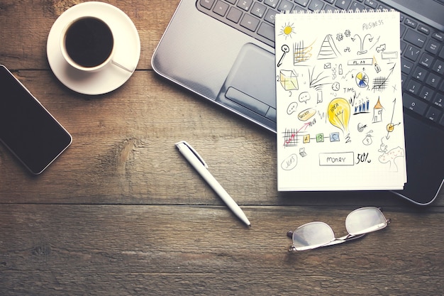 Notepad over laptop, coffee, writing instruments and coffee on wooden table