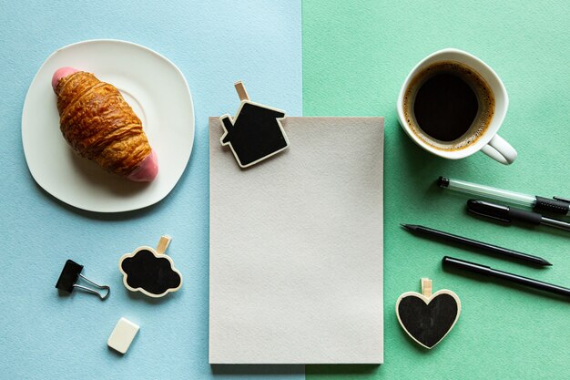 Blocco note, matita, croissant e caffè su una scrivania verde-blu.