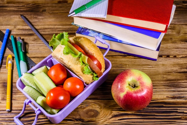 Notepad pens ripe apple stack of books and lunch box with hamburger cucumbers and tomatoes on rustic wooden table