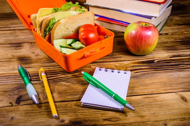 Foto blocco note penne mela matura pila di libri e pranzo al sacco con panini cetrioli e pomodori su tavola di legno rustico