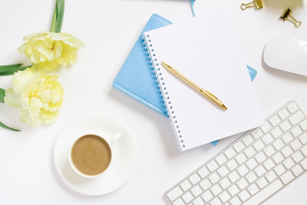 Notepad with a gold pen yellow tulip flowers a cup of coffee a keyboard and a cup of coffee on a white background Stylish workplace of a blogger or freelancer