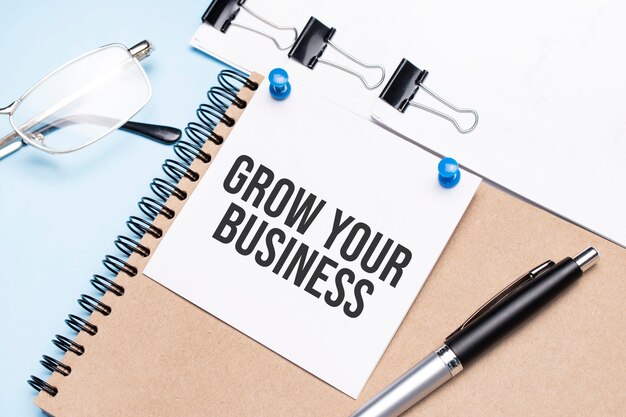 안경, 메모장 및 클립이있는 종이가있는 사무실 테이블에 GROW YOUR BUSINESS 텍스트가있는 메모장.