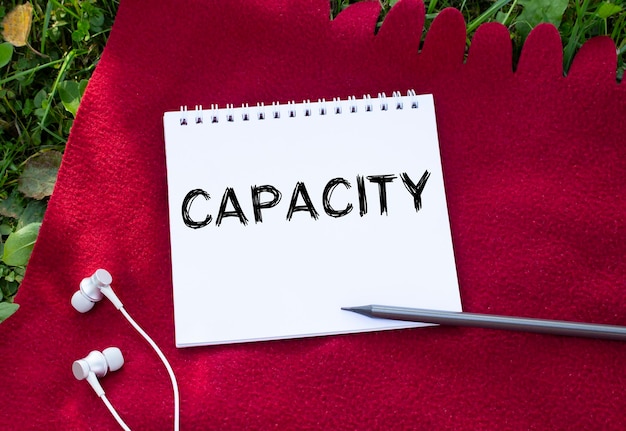 CAPACITYの碑文が書かれたメモ帳ヘッドフォンが近くにあります背景には赤い格子縞と緑の草があります