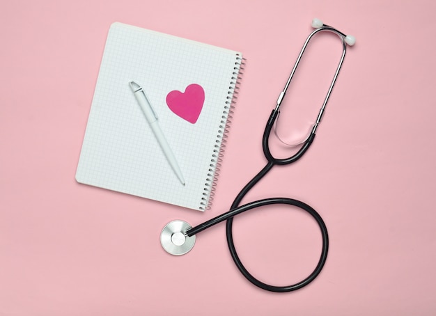 Blocco note con la penna, cuore decorativo, stetoscopio su fondo pastello rosa, concetto medico, vista superiore, tendenza di minimalismo