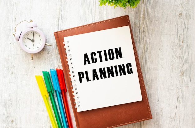 Photo notepad with the text action planning on a wooden table. brown diary and pens.