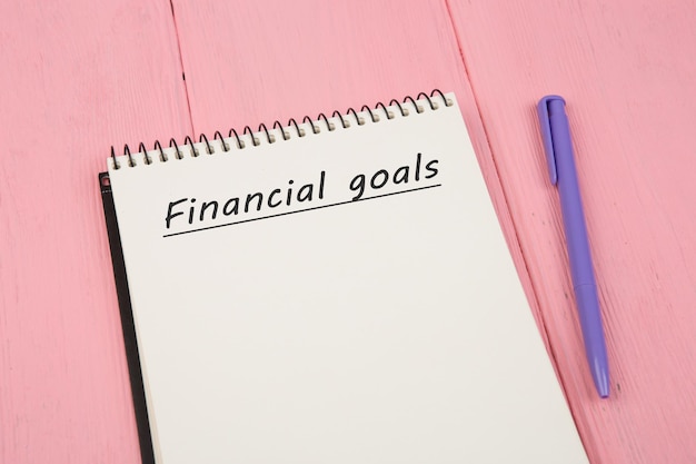 Блокнот с текстом Финансовые цели и ручка на розовом деревянном столе