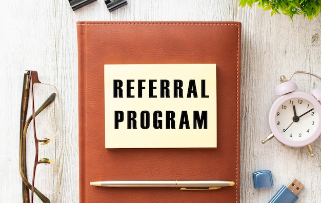 木製のテーブルに「REFERRALPROGRAM」というテキストが書かれたメモ帳。茶色の日記とペン。