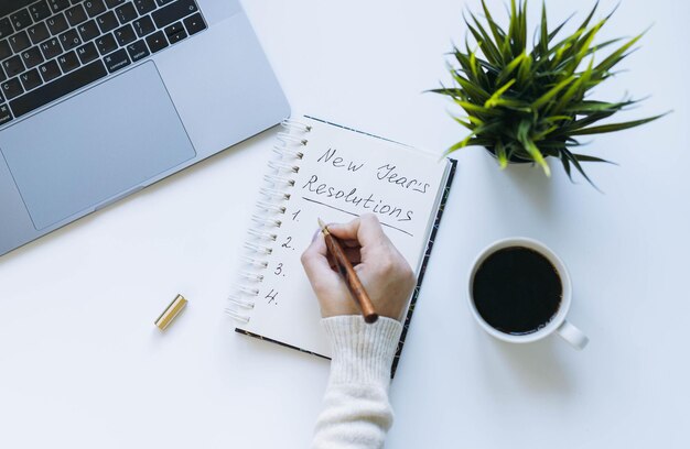 Notepad and woman hand writing new year resolutions Flower laptop and coffee cup on background