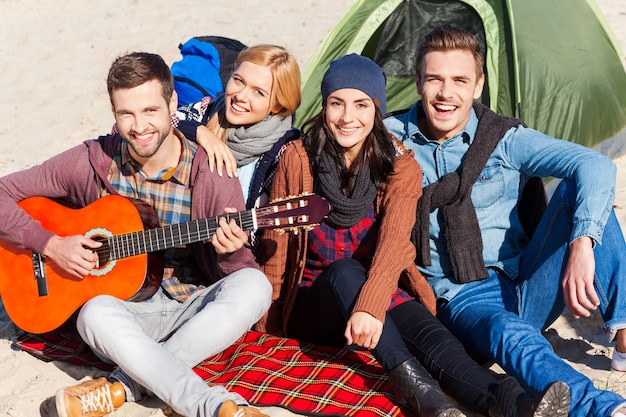 Только лучшие друзья. Вид сверху четырех молодых счастливых людей, сидящих вместе возле палатки, пока молодой красивый мужчина играет на гитаре и улыбается
