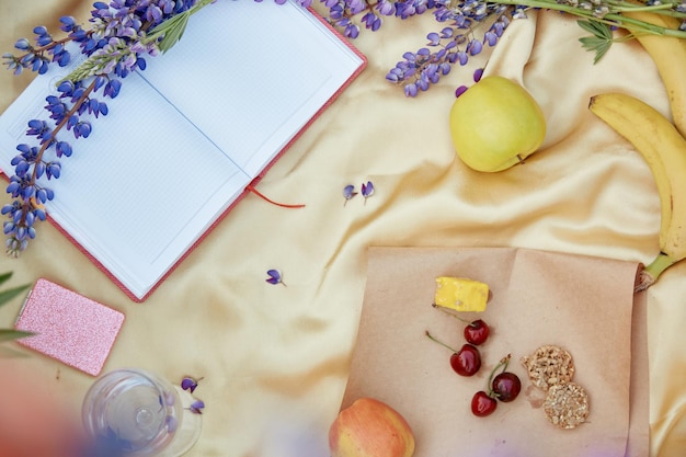 Notitieblok openen mock up met wijnglas lupine paarse bloemen appel bananen kersen halva cookies op gouden achtergrond Zomerpicknick plaats voor tekst Bovenaanzicht Nabijheid van de natuur zelfontdekking concept