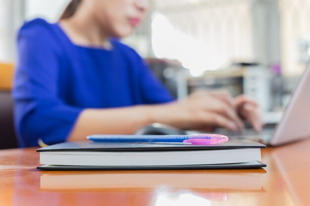 Notitieboekje en pen op de lijst met vrouw die op laptop op onduidelijk beeldachtergrond woking.