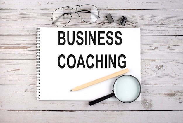 Notitieboekje met tekst business coaching op de houten tafel met pen, vergrootglas en bril