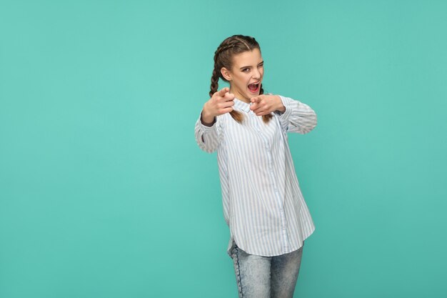 Now is your time. portrait of funny beautiful girl in blue striped t-shirt and pigtail hair pointing an looking at camera with open mouth and winking. indoor studio shot isolated on green background.