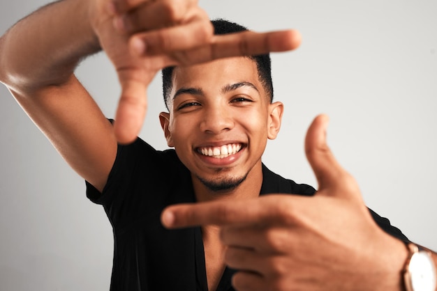 Now stand perfectly still Portrait of a cheerful young man making a frame with his fingers while standing against a grey background inside of a studio