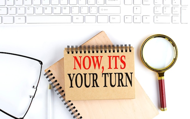 NOWIT'S YOUR TURN text in the office notebook with keyboard magnifier and glasses business concept