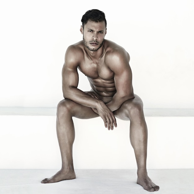 Nude body and man in portrait isolated on a wall background for health fitness and wellness Naked strong and face of a sexy male model sitting with muscles for power sports and exercise results