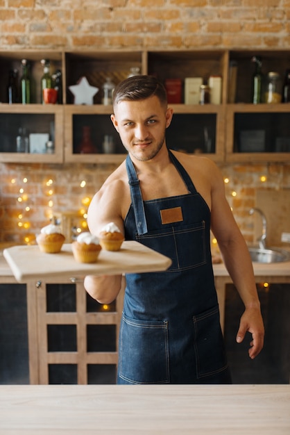 Nude husband in apron holds tray with sweet dessert on the kitchen. Naked male person preparing breakfast at home, food preparation without clothes