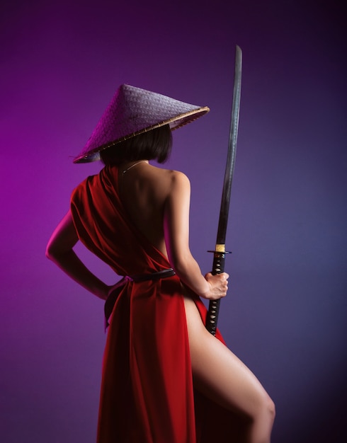 The nude slender Asian woman in a red cape and an Asian hat with a katana in her hand image of a samurai on a neon background