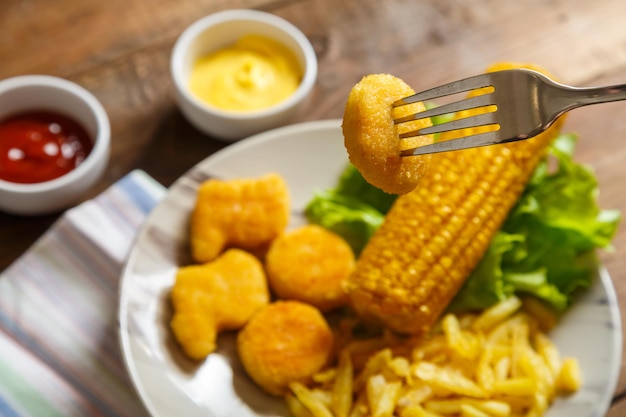 Наггетсы, картофель фри и кукуруза в початках на листьях салата на тарелке с наггетсами на проколотой вилке. Горизонтальное фото