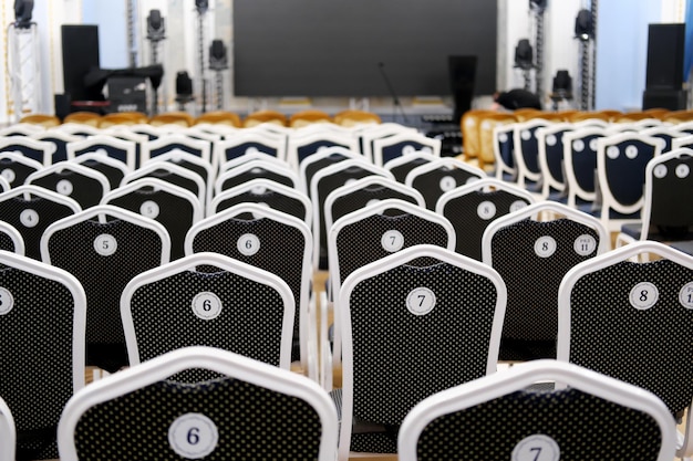 Numbered chairs stand in a row against the backdrop of a large screen stage