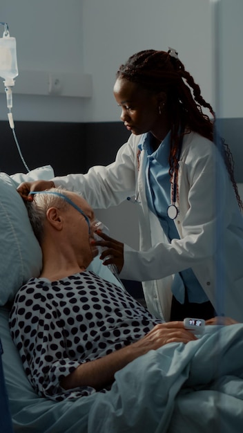 간호사와 아프리카계 미국인 의료진은 병원 병동 침대에 있는 환자를 위해 산소 튜브와 맥박 모니터를 사용합니다. 수술에서 회복하기 위해 소생법이 필요한 IV 드립백을 든 노인