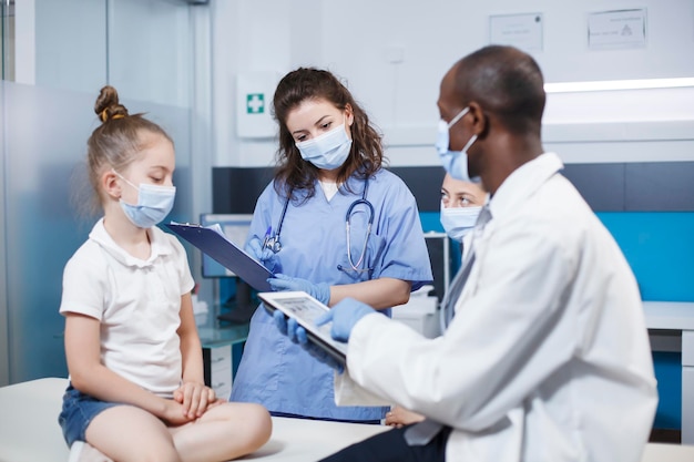 Nurse in blue scrubs filling out a clipboard with information on the girl Face masks were worn by the patient her mother and the multicultural medical personnel An atmosphere of professionalism