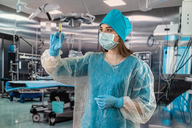 Nurse in a blue uniform with a mask and gloves behind a hat holding a syringe with medicine