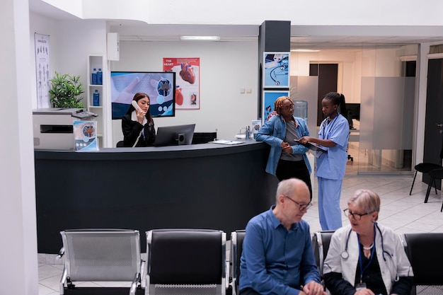 Nurse consulting patient at facility reception counter, analyzing report forms and checkup papers to give treatment against disease diagnosis. Specialist helping woman with recovery in waiting area.