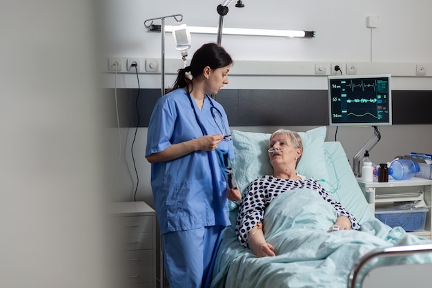 Nurse discussing with elderly patient laying in hospital bed during visit about chest x-ray