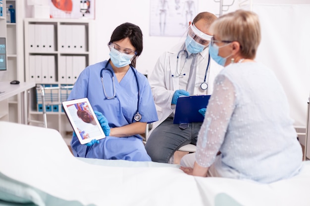 Nurse explaining heart diagnosis in cardiology to senior woman on tablet pc in time of covid pandemic