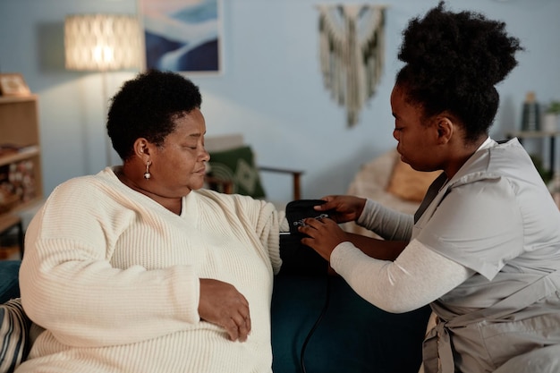 nurse fastening cuff of tonometer on elderly black woman checking blood pressure at home