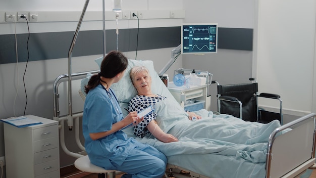 Nurse giving assistance to senior patient with disease in bed. Medical assistant and doctor doing healthcare checkup for pensioner with oxygen tube and IV drip bag in hospital ward.