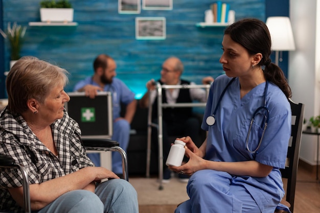 Nurse giving new medication treatment to senior female resident in common room of nursing home. Male nurse attending to elderly male patient, seated on sofa in background.