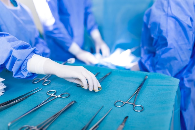 Nurse hand taking surgical instrument for group of surgeons at background operating patient in surgical theatre Steel medical instruments ready to be used Surgery and emergency concept