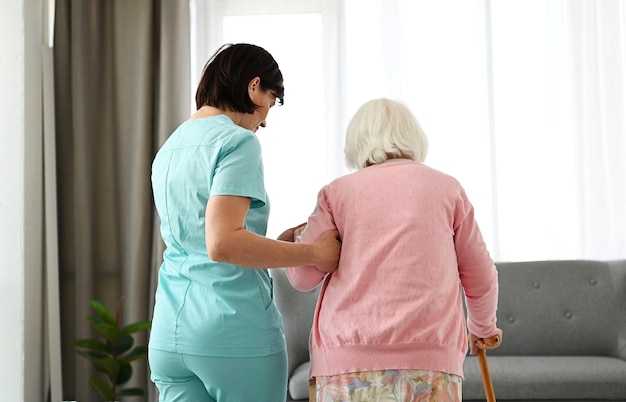간호사는 노인 여성이 방을 돌아다니는 것을 돕습니다. 노인에 대한 돌봄과 지원의 개념을 구현합니다.