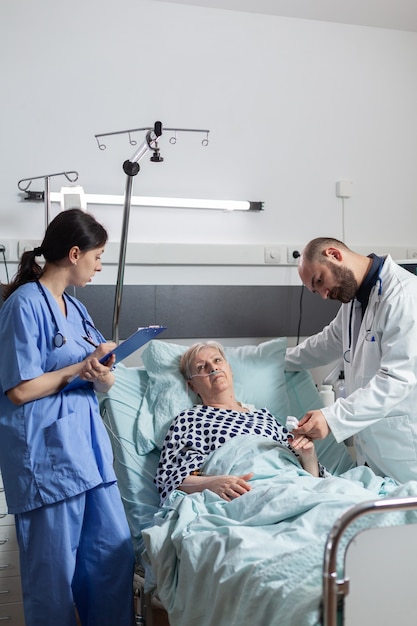 Nurse is taking notes on clipboard during consultation of senior woman, while doctor is reading data from oxymeter about pulse, blood pulse and oxygen saturation attached on sick patient attached on f