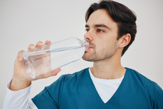 病院の白い背景に隔離されたスタジオでボトルに入った水を飲む看護師男性と健康ウェルネスまたは体の栄養医療専門家の水分補給と休憩中ののどが渇いた外科医の液体