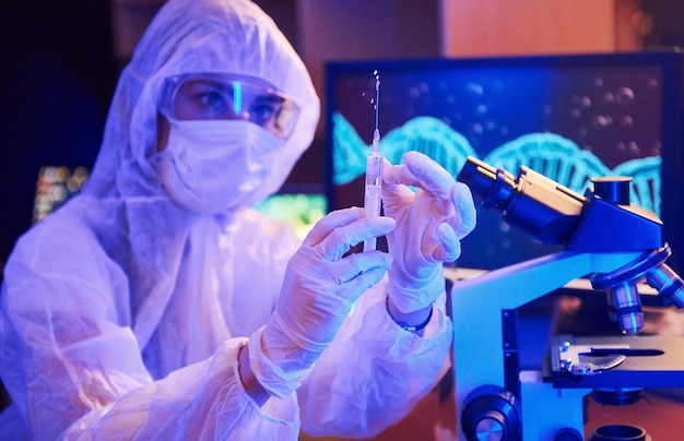 Nurse in mask and white uniform holding syringe and sitting in neon lighted laboratory with computer and medical equipment searching for Coronavirus vaccine