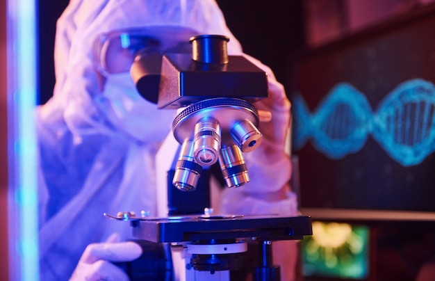 Nurse in mask and white uniform sitting in neon lighted laboratory with computer, microscope and medical equipment searching for Coronavirus vaccine