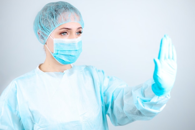 Медсестра в медицинском халате, маске и защитных перчатках стоит боком, протянув руку в жесте «стоп».
