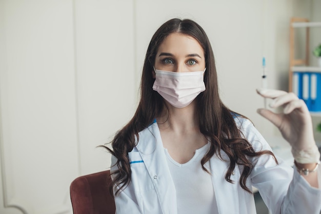 Nurse posing with syringe for vaccine injection wearing white robe rubber gloves and medical mask co...