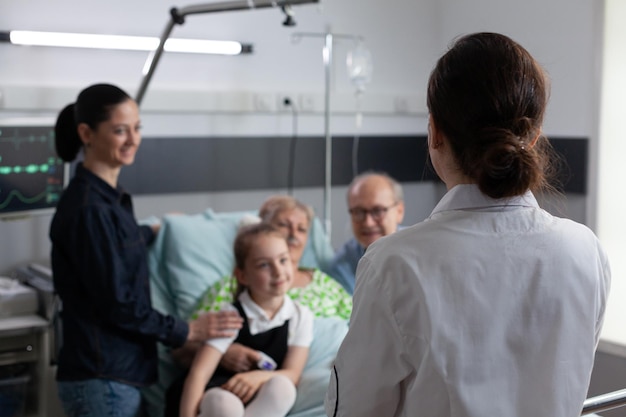 Nurse practitioner visiting happy elderly patient room in\
recovery. sick grandmother smiling family chatting with\
unidentified female geriatric professional doctor at medical clinic\
room.