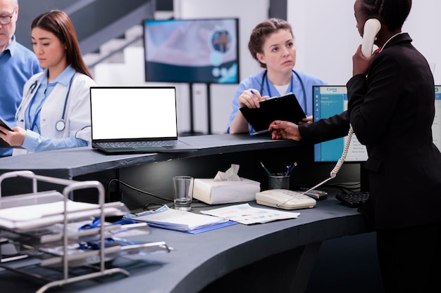 病院の待合室での診察中に病気の症状について話し合う患者の治療に従事する看護師と受付係。カウンターの机の上に立っている白い画面のラップトップ コンピューター