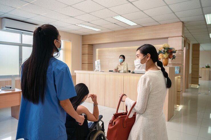 Photo 간호사 서비스가 휠체어에 탄 아시아 환자 여성을 병원에 입원시키기 위해 접수원에게 연락하도록 강요합니다.