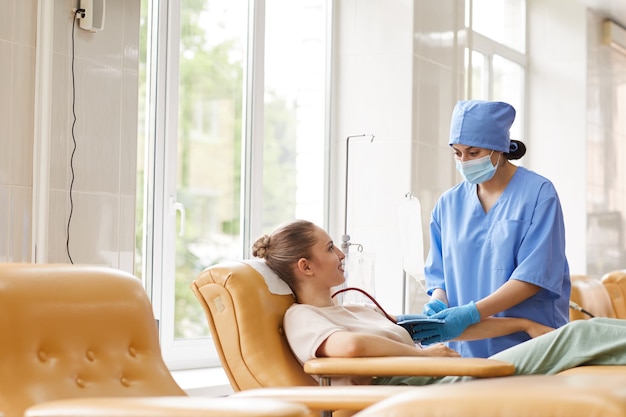 Nurse in uniform taking blood from donor while she lying on the couch in the laboratory