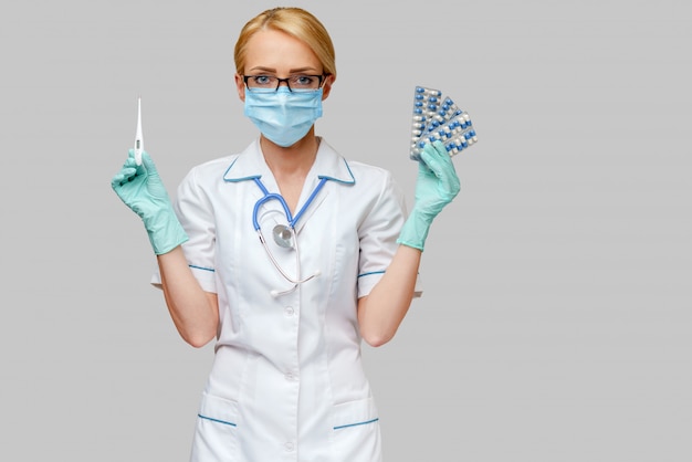 Nurse wearing protective mask and latex gloves and holding blisters of pills and thermometer