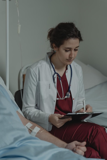 A nurse in a white coat sits next to a patient in a hospital bed.