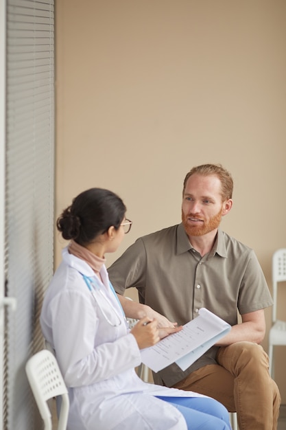 Nurse in white coat sitting with medical card and consulting patient at hospital