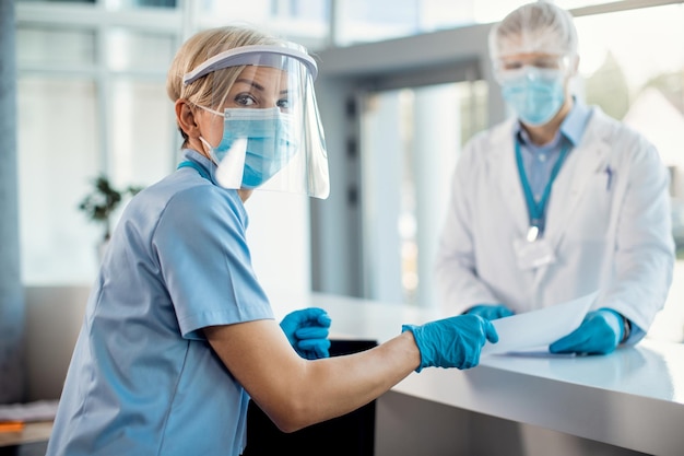 Nurse with face shield and mask working at reception desk at the hospital